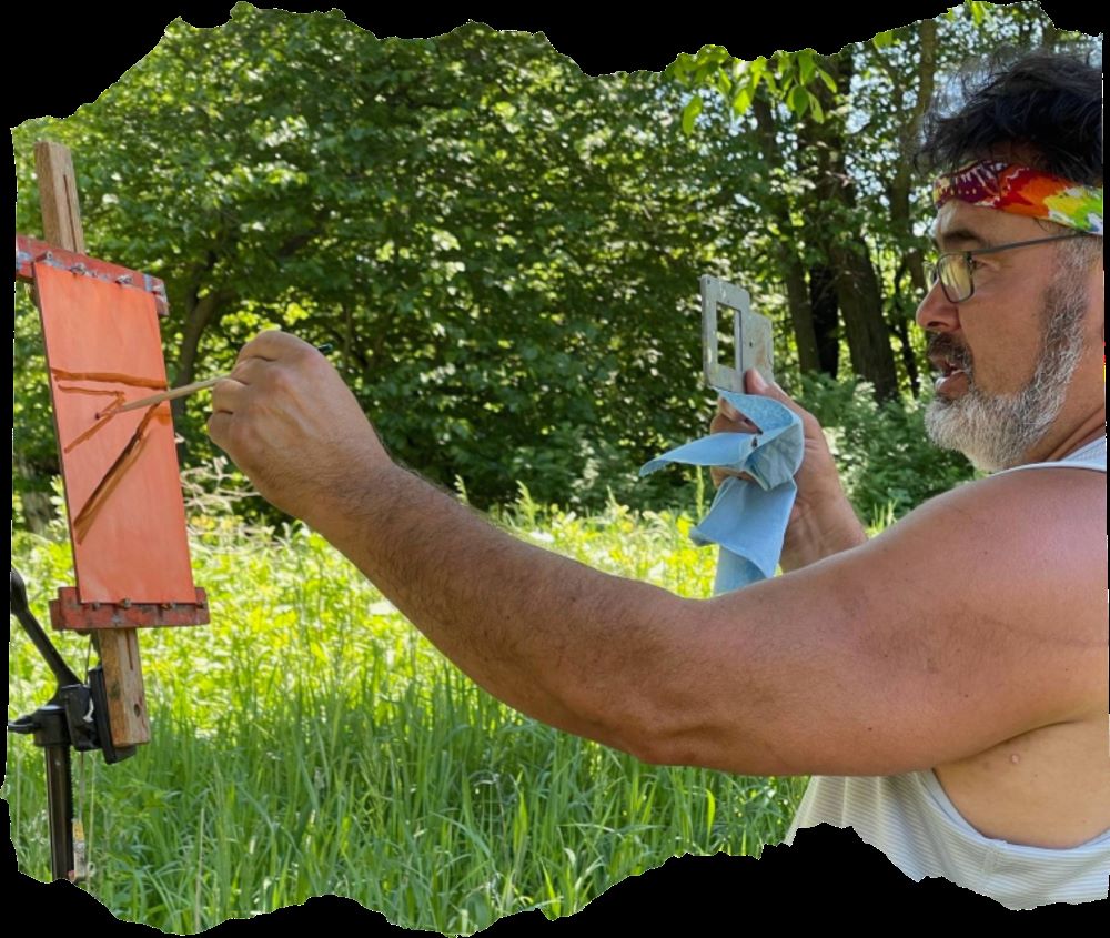 A man sits outdoors among tall grasses and trees, painting on a small canvas on an easel.