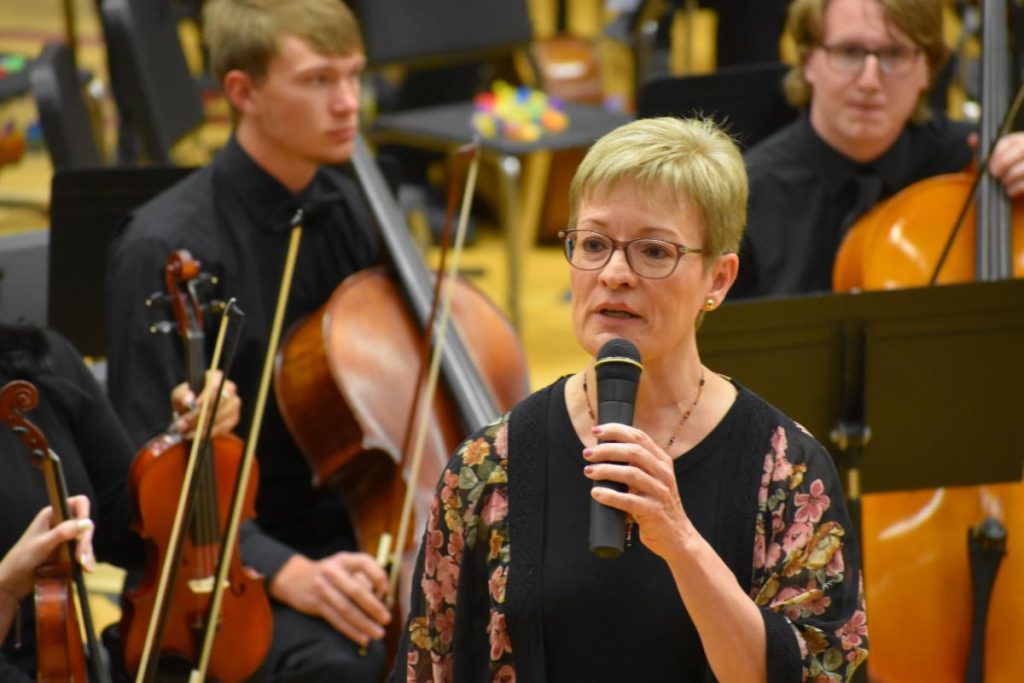 A woman standing in front of young violin and cello players speaks into a microphone.