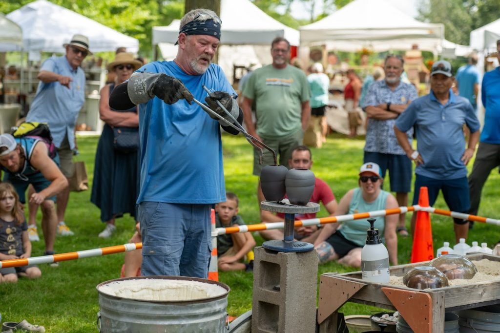 A man uses large tongs to pick up a hot vase while adults and children watch behind him.