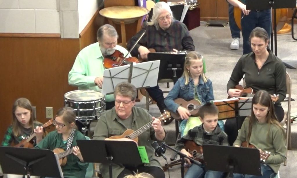 A group of seated children and adults play ukeleles and fiddles.
